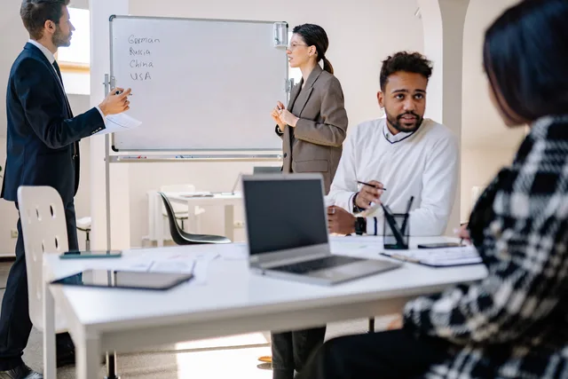 A diverse group of professionals engaged in a collaborative office meeting with laptops and a whiteboard. Where AI actually helps B2B social selling, from account research to assurance